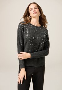 Dunkelgrauer gestrickter Pullover mit rundem Halsausschnitt, verziert mit verstreuten silbernen Pailletten und gerippten Bündchen. Kombiniert mit schwarzen Hosen, die Verzierungen aufweisen.