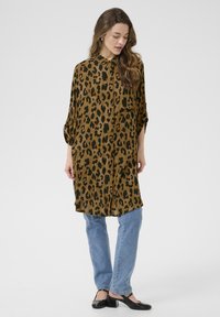 Robe chemise en imprimé léopard brun et noir, avec un design à boutons sur le devant, ourlet arrondi, associée à un jean bleu clair et des ballerines noires.