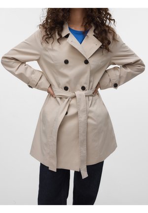 Kobieta w beżowym płaszczu trenczu dwurzędowym z czarnymi guzikami i paskiem związanym w talii, niebieską bluzką i ciemnymi spodniami, z rękami na biodrach na białym tle.