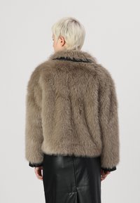 Faux fur jas in lichtbruin met een zachte, pluizige textuur, voorzien van een ronde kraag en zwart leren accenten langs de randen.