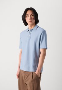 Himmelblaues Polohemd mit kurzen Ärmeln und strukturiertem Finish, Drei-Knopf-Leiste und kontrastierendem dunkel gefärbtem Kragen. Getragen mit beigen Hosen.