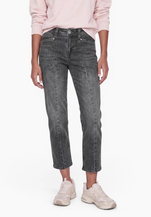 Person iført falmede sorte korte jeans, en lyserød langærmet skjorte og hvide sneakers, der står mod en hvid baggrund.