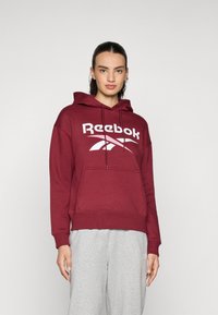 Felpa con cappuccio bordeaux con un grande logo bianco Reebok, tasca a marsupio e polsini a costine, abbinata a pantaloni della tuta grigi.