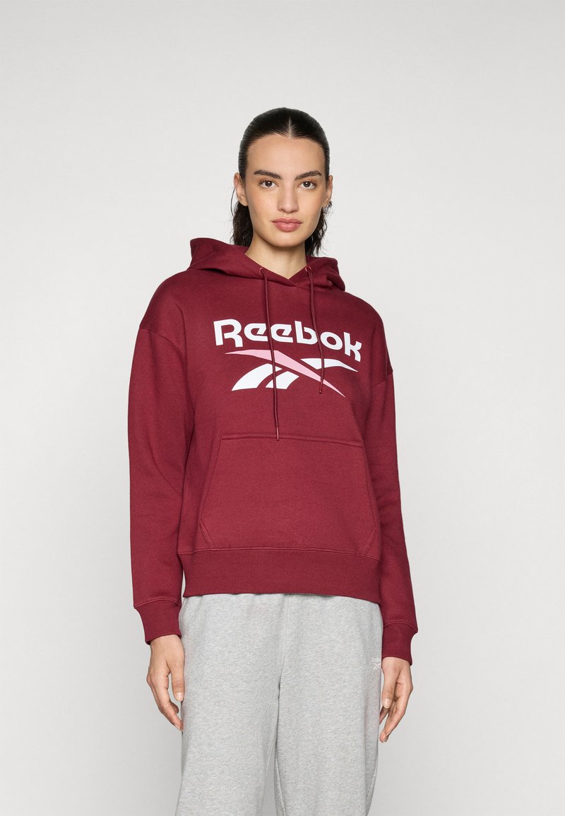 Felpa con cappuccio bordeaux con un grande logo bianco Reebok, tasca a marsupio e polsini a costine, abbinata a pantaloni della tuta grigi.