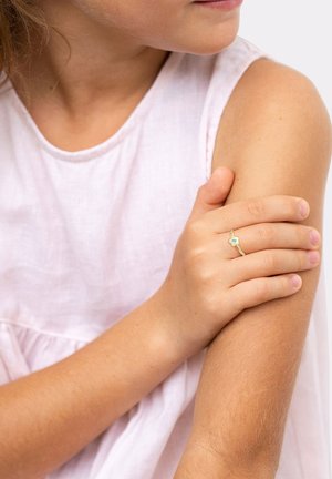 Bague en or avec un détail de fleur bleu clair portée sur la main d'un enfant, peau lisse, sur un fond de tissu rose pâle.