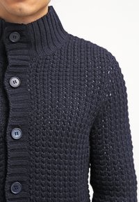 Muž má na sobě cardigan z texturovaného pleteného materiálu v tmavě námořnické modré barvě s žebrovaným stojáčkem a se čtyřmi viditelnými černými knoflíky zapnutými.