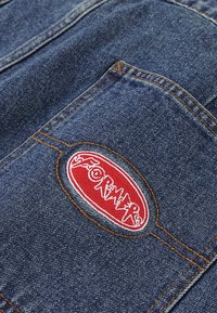 Denim fabric in dark blue with orange stitching. Features a circular red embroidered patch with white lettering reading "FORMERS."
