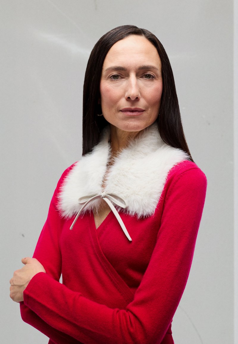 Hosszú sötét hajú nő piros megkötős pulóverben és fehér műszőrme gallérral, szalaggal megkötve, áll egy semleges háttér előtt.