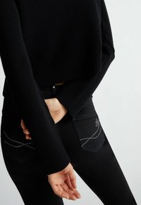 Maglione cropped nero a coste con maniche lunghe e jeans neri con borchie decorative argentate sui taschini posteriori.