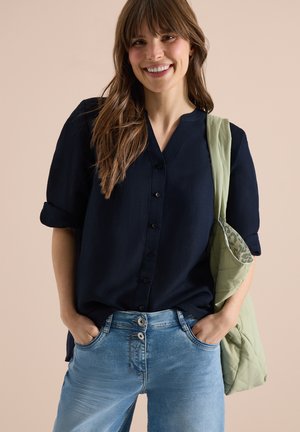 Junge Frau lächelt mit langen braunen Haaren, trägt ein schwarzes Hemd mit Knöpfen, blaue Jeans und eine hellgrüne gesteppte Schultertasche.