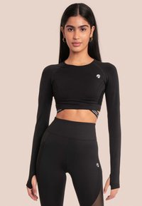 Zwarte longsleeve crop top met een aansluitend ontwerp, subtiele mesh-details en logo; gecombineerd met hoge zwarte leggings met uitsnijdingen.