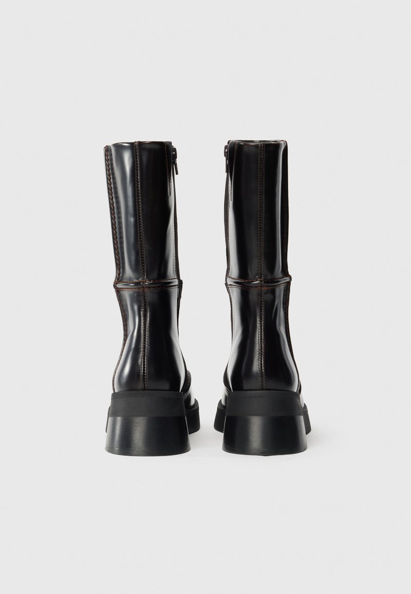 Bottes en cuir noir montantes avec une finition brillante, dotées d'une semelle épaisse noire et de coutures marron contrastantes le long des coutures latérales.