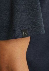 Lähikuva tummansinisestä kankaasta valmistetusta hihasta, jossa on pieni musta etiketti, jossa on valkoinen minimalistinen kaarisuunnittelu henkilön käsivarressa.