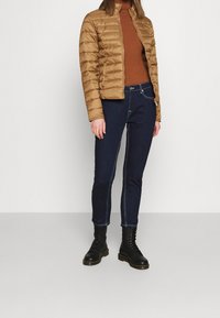 Brun dunjakke, brun ribbet rullekrave, mørk denimjeans med hvide syninger og sorte snørebånds-kampstøvler. Stylet med en tætsiddende silhuet.