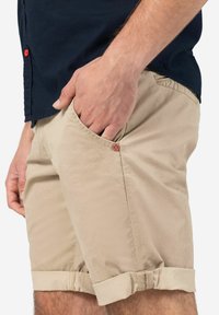 Beige cotton shorts with rolled cuffs, featuring a red logo detail on the front pocket and side seam pockets. Soft texture and casual design.
