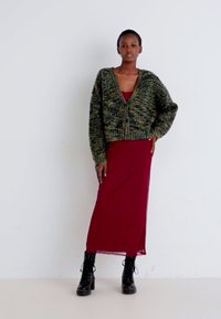 Cardigan oversize lavorato a maglia in tonalità di verde e giallo, abbinato a un vestito aderente color bordeaux. L'outfit è completato da stivali neri con lacci.