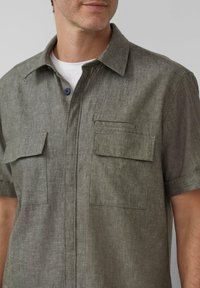 Korte mouwen blouse in olijfgroen stof met een gestructureerde afwerking, voorzien van twee borstzakken en een donker knoopdetail.
