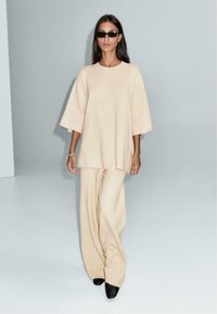 Beżowy oversize sweter z opuszczonymi ramionami w zestawie z szerokimi spodniami o dopasowanym kroju. Gładka faktura; modelka nosząca ciemne okulary przeciwsłoneczne.