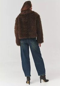 Brązowa kurtka z sztucznego futra o skróconym fasonie, z wysokim kołnierzem i długimi rękawami, zestawiona z niebieskimi jeansami o wysokim stanie. Widoczne czarne botki za kostkę.