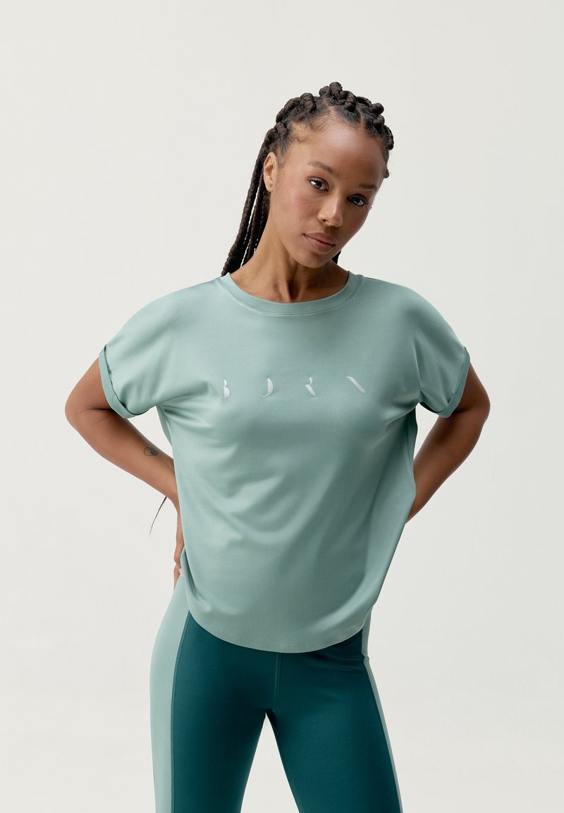 Voľný, krátky rukáv tričko v jemnej mätovej zelenej farbe s hladkou textúrou. Má biele logo na prednej strane, skombinované s ladnými legínami.