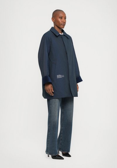 Navy blue lightweight jacket with a collar, side pockets, and contrasting cuffs. Worn over relaxed-fit denim jeans and black heeled shoes.