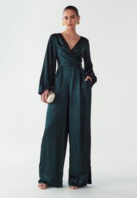 Smaragdgrüner Overall mit Wickeldesign, langen Ballonärmeln, Seitentaschen und einer glatten, reflektierenden Textur. Kombiniert mit einer kleinen goldenen Tasche.