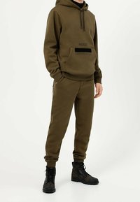 Oliwkowa bluza z kapturem z przednią kieszenią i czarnym logo, w połączeniu z dopasowanymi spodniami zwężającymi się ku dołowi. Czarne buty za kostkę z detalami teksturalnymi.