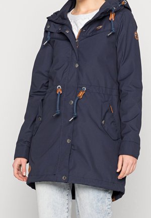 Femme portant une parka à capuche bleu marine avec cordons de serrage et boutons-pression, associée à un jean bleu clair.
