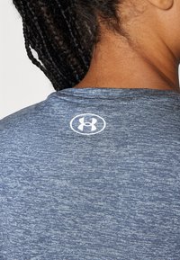 Kortermet blå sportskjorte med teksturert stoff og en hvit Under Armour-logo på baksiden av halsen.