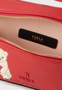 Κόκκινη τσάντα από υφή δέρματος με μπεζ εσωτερικό, χρυστό φερμουάρ και μοτίβο αρκούδας. Μαύρη ετικέτα με την επιγραφή "FURLA SINCE 1927."