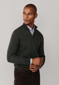 Dunkelgrüner Reißverschluss-Pullover aus strukturiertem Stoff über einem hellblauen Hemd mit Kragen, kombiniert mit braunen Cordhosen.