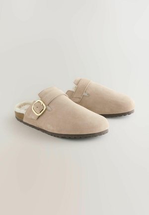 Chaussures slip-on en suède beige clair avec un intérieur doux, une sangle réglable et une boucle dorée, dotées d'une semelle en caoutchouc texturée.