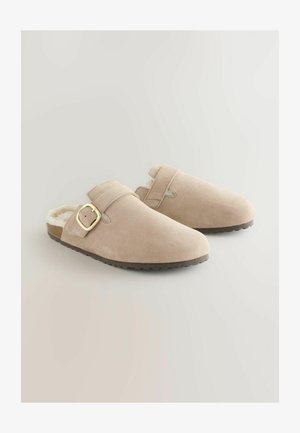 Chaussures slip-on en suède beige clair avec un intérieur doux, une sangle réglable et une boucle dorée, dotées d'une semelle en caoutchouc texturée.