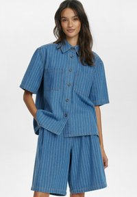 Blå denimkortärmad skjorta och matchande shorts med vita vertikala ränder. Har två bröstfickor och knappstängning framtill.