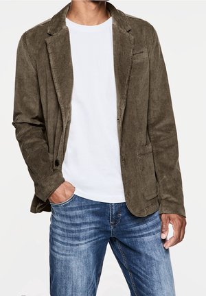 Uomo che indossa un blazer di velluto a coste verde oliva, t-shirt bianca e jeans blu, con una mano nella tasca, su sfondo bianco.