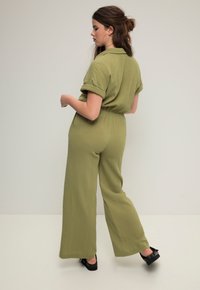 Olivengrüner, strukturierter Jumpsuit mit kurzen umgeschlagenen Ärmeln und tailliertem Gürtel; weite Hosenbeine; getragen mit schwarzen Plateausandalen.