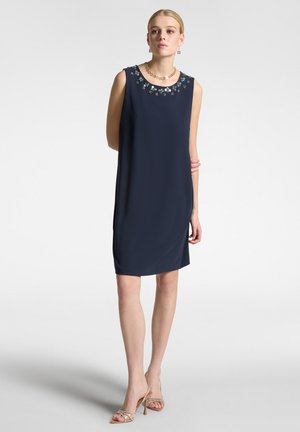 Abito senza maniche blu navy con ricami floreali intorno allo scollo, texture liscia e lunghezza al ginocchio. Abbinato a sandali con tacco nudo.