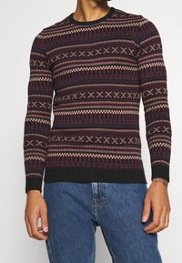 Pull en tricot avec un motif géométrique en rouge foncé, crème et bleu marine, doté d'un col rond côtelé et de poignets côtelés. Coupe standard et surface texturée.