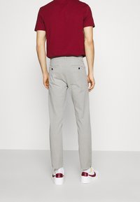 Homme portant un pantalon sur mesure gris clair, des baskets Nike blanches avec un swoosh rouge, et une chemise à manches courtes rouge foncé rentrée, se tenant de dos.
