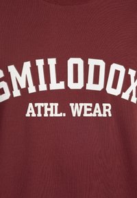 Bordeaux sweatshirt gemaakt van zacht materiaal met groot wit tekst "SMILODON" en "ATHL. WEAR." Ribgebreide textuur met een klassiek ronde hals ontwerp.