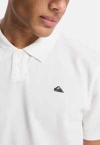 Λευκό πόλο πουκάμισο με κλασικό γιακά, πατιλέτα τριών κουμπιών και μαύρο κεντητό λογότυπο στο αριστερό στήθος. Υφασμα από βαμβάκι με υφή.