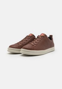 Brown leather sneakers with a textured finish, featuring a light-colored sole, perforated toe, and a pull tab on the heel. Red logo detail.