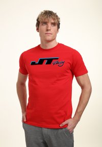 Červené bavlněné tričko s černým a modrým logem "JT Racing" na přední straně. Krátké rukávy a kulatý výstřih, kombinované se šedými kalhotami.