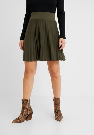 Frau trägt einen olivgrünen, plissierten Rock, ein schwarzes Langarmoberteil und braune knöchelhohe Stiefel mit Schlangenledermuster, steht vor einem einfarbigen Hintergrund.