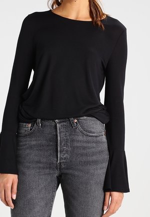 Mujer vistiendo una blusa negra de manga larga con puños acampanados y jeans negros de cintura alta desgastados, de pie contra un fondo sencillo.