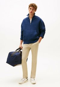 Felpa zip in blu navy con motivo geometrico testurizzato, pantaloni chinos beige e sneakers bianche, mentre tiene una borsa da viaggio blu scuro con dettagli marroni.