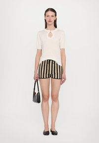 Kratkoročni kremni top z detajlom v obliki ključavnice na vratu, vparen z black and cream patterned šortkami. Drži majhno črno torbico; ravne čevlje.