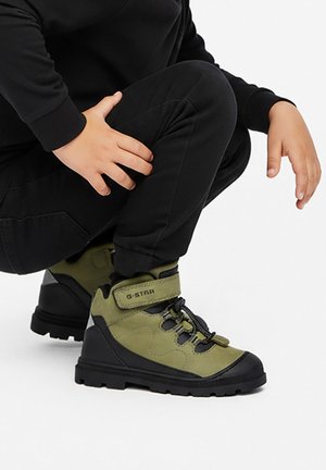 Bambino con pantaloni neri accovacciato, che mostra scarpe alte G-Star verdi e nere con cinturini in Velcro e lacci su sfondo bianco.