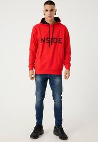 Sudadera roja con capucha que presenta el texto "INSIDE" en negro, con un bolsillo frontal, combinada con jeans de mezclilla azul y zapatillas negras.
