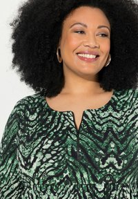 Vihreä kuviollinen mekko pyöreällä kaula-aukolla ja edessä olevan vetoketjun kanssa. Kangas esittää mustan ja vihreän käärmemäisen kuvion rakennetta.